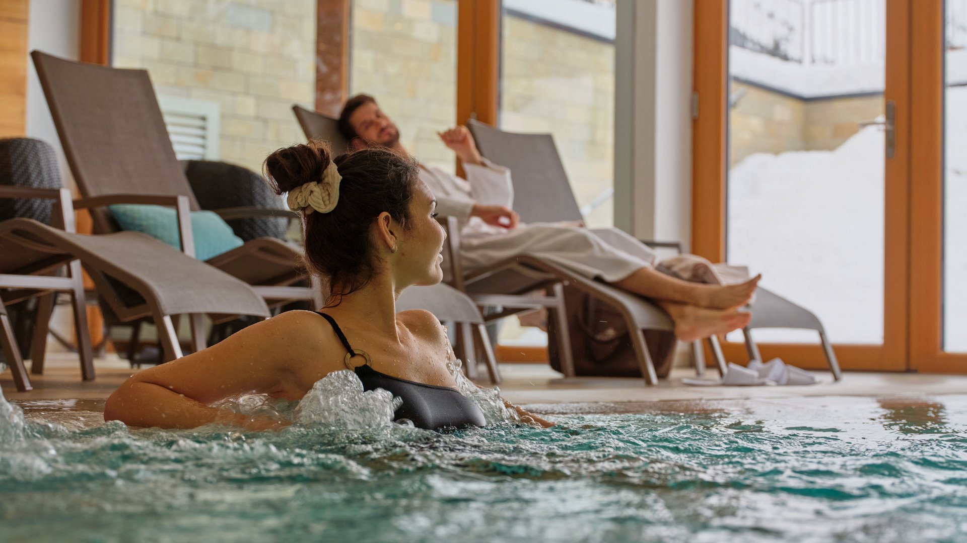 Frau im Whirlpool und Mann entspannt auf Liegestuhl in Spa mit Schneeblick