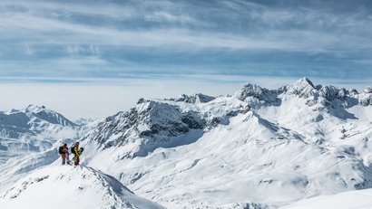 Zwei Snowboarder stehen auf schneebedecktem Gipfel mit Alpen im Hintergrund