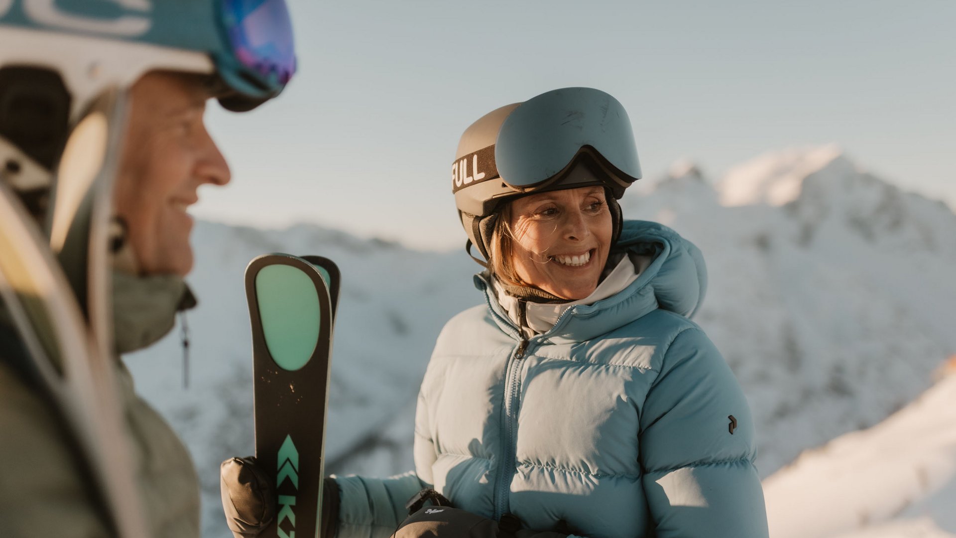 Frau in blauer Skijacke und Helm hält Ski in verschneiter Berglandschaft