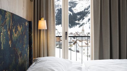 Bett und Lampe vor Fenster mit Blick auf verschneites Dorf und Berge