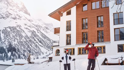Zwei Skifahrer vor einem Haus im Schnee mit Berg im Hintergrund