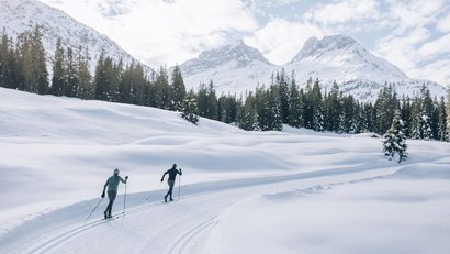Zwei Langläufer auf verschneiter Strecke vor Tannen und Bergen