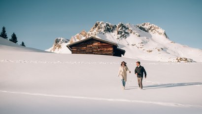 Paar spaziert im Schnee vor einer Holzhütte und verschneiten Bergen