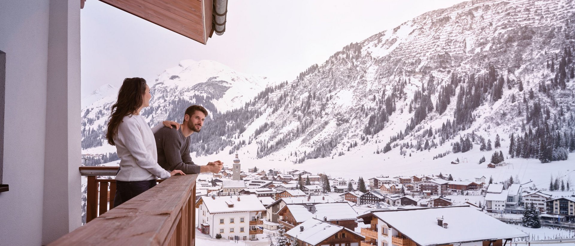 Paar auf Balkon blickt auf verschneites Dorf und Berge